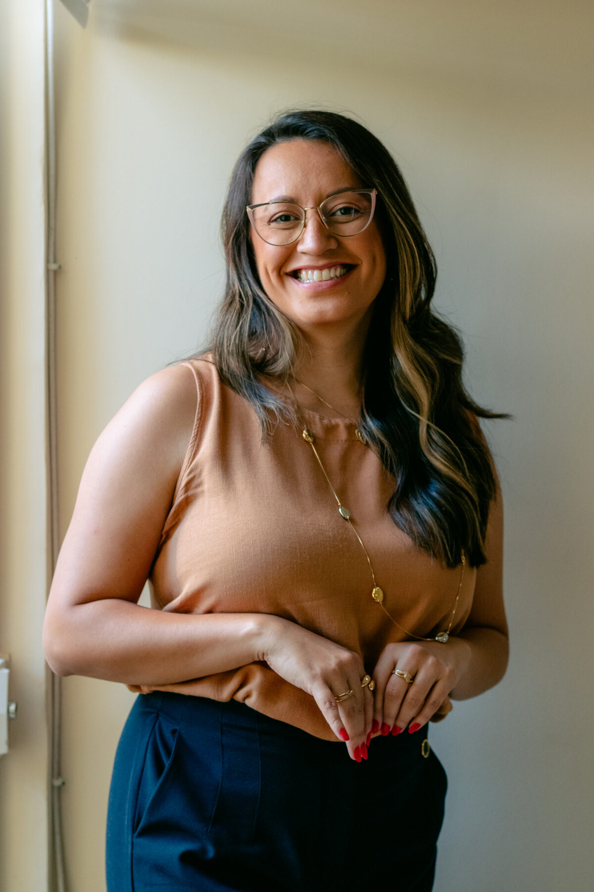 Amanda Moura em pé, sorrindo para a câmera, com blusa areia sobre fundo creme