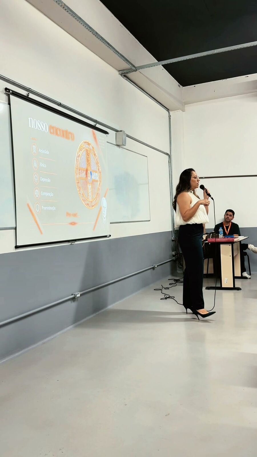 Amanda Moura palestrando ao microfone, com o telão da apresentação ao fundo